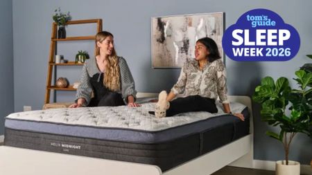 The image shows a two female mattress testers sat on a white and navy blue mattress, which is placed on a white bed frame in a neutrally decorated bedroom. Overlaid on top is a blue Tom's Guide Sleep Week 2026 badge overlaid in the top right corner.