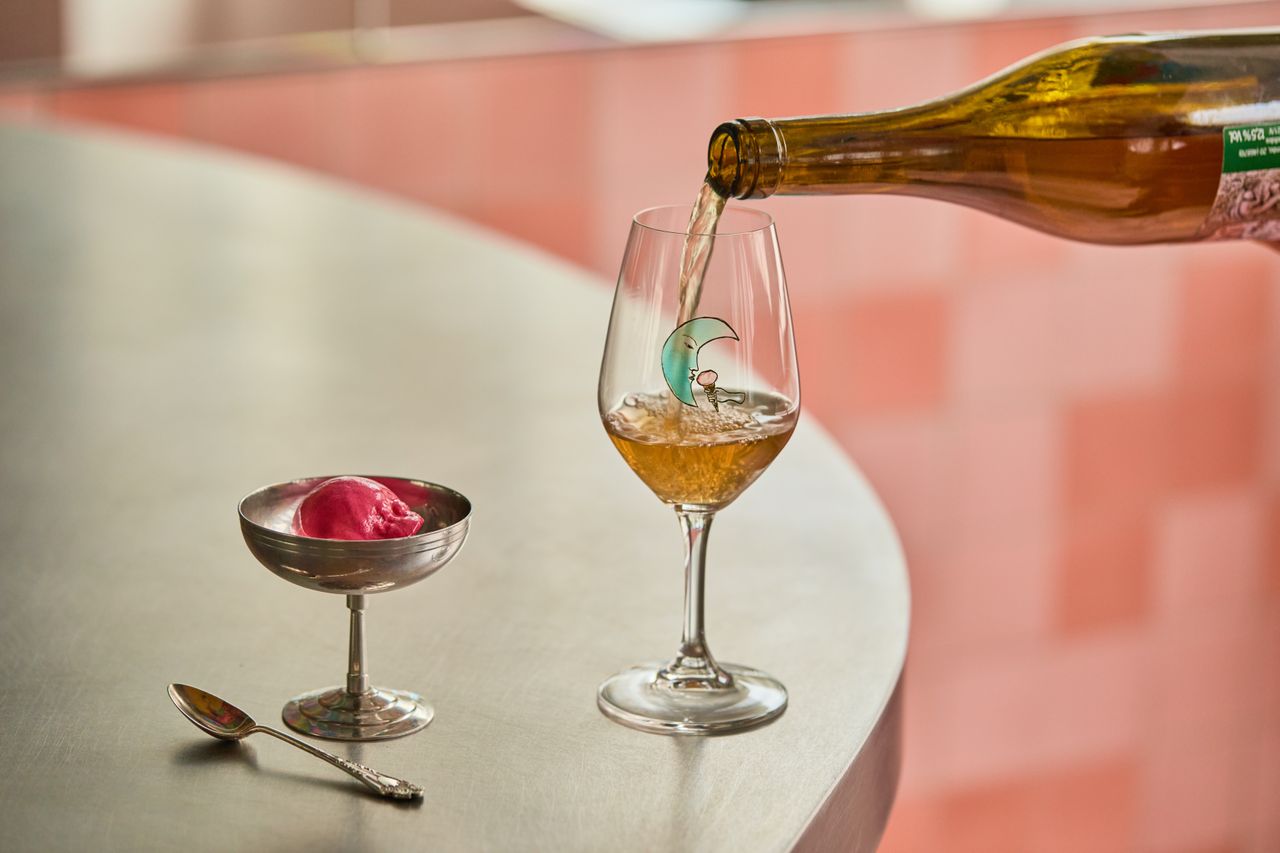 A glass of wine with a light blue moon motif is being filled up by a person standing on the left of a chrome table, and sits on it next to a chrome ice cup with a pink scoop in it, and a chrome spoon.