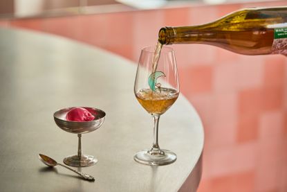 A glass of wine with a light blue moon motif is being filled up by a person standing on the left of a chrome table, and sits on it next to a chrome ice cup with a pink scoop in it, and a chrome spoon.