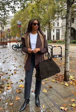 how to wear chelsea boots is shown in a photo of a woman on the sidewalk wearing a brown blazer over a white t-shirt with black skinny jeans, a brown suede bag, black heeled chelsea boots, and a tan cardigan