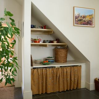 An alcove under the stairs with open shelving, a storage basket and the bottom covered with a curtain