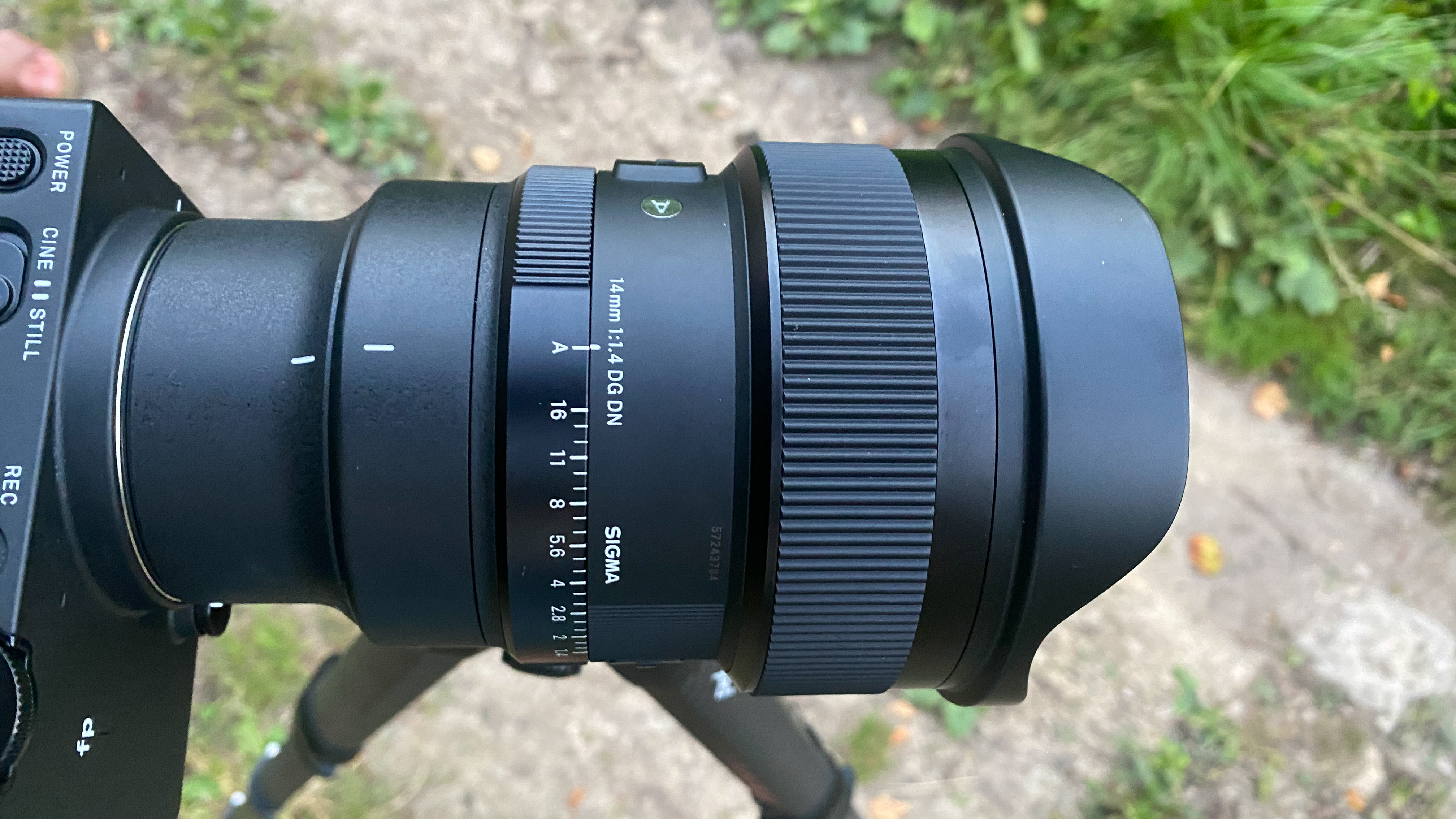 A top-down view of a black camera lens with two ridged adjustment rings, which is mounted to a tripod and attached to a rectangular black camera, half cut out of frame. The lens body obscures the tripod, except for two legs telescoping toward the ground.