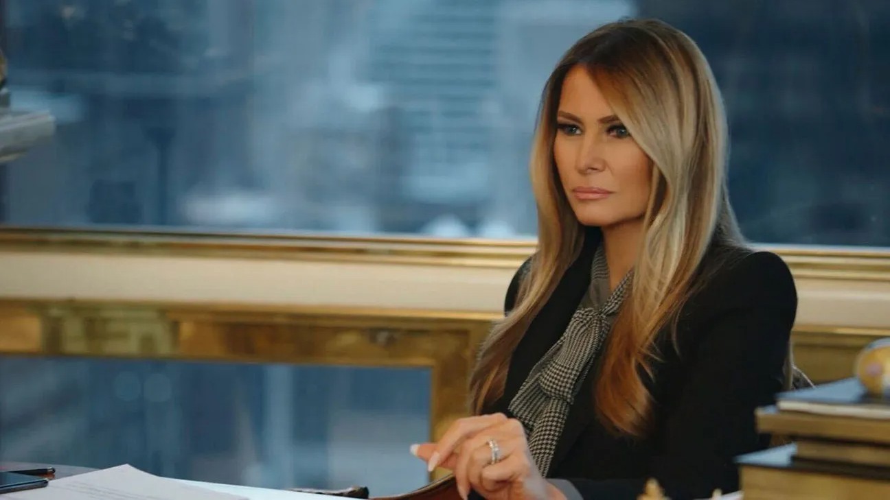 Melania Trump sits at a desk in a professional setting, wearing a black blazer over a patterned pussy-bow blouse.