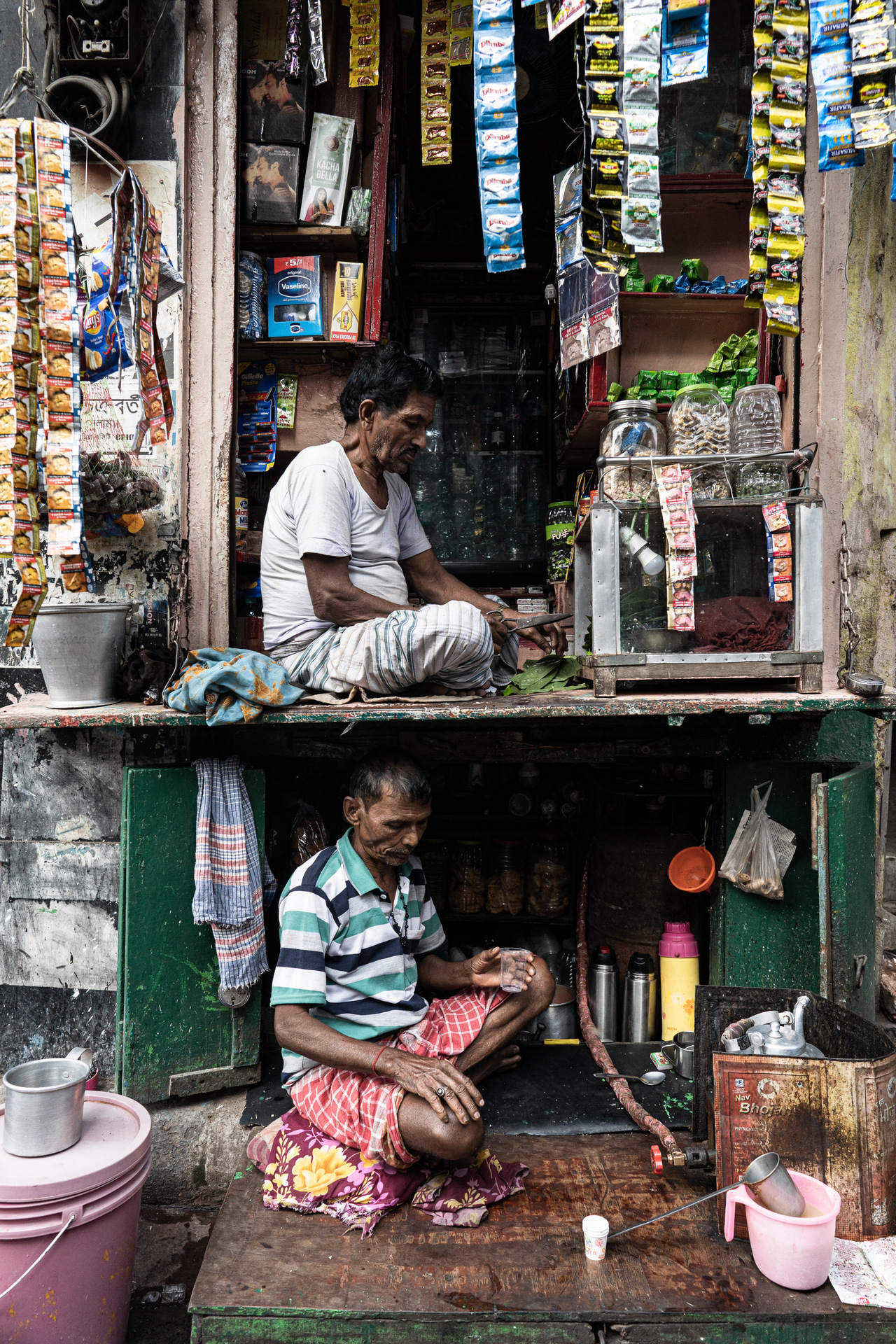 Two men sit in a bustling storefront filled with snacks and beverages, surrounded by colorful packaging hanging on display