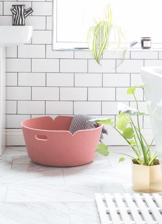 A pink washing basket with grey towel in a white tiled bathroom and plant - Muuto Restore Round BaSket