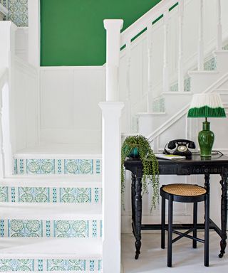 Hallway with white walls and painted paneling, green painted wall beside staircase, wallpapered stairs, white flooring, dark wood console table with green table lamp, plant and black telephone, black wood and rattan stool