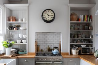 kitchen with grey painted Shaker units, wood worktop, metro tiles in cooker alcove, and open shelving either side of the cooker filled with jars, crockery and cookbooks