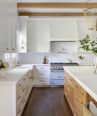 A white kitchen with a wooden island, exposed ceiling beams, gold hardware, and a light blue range cooker