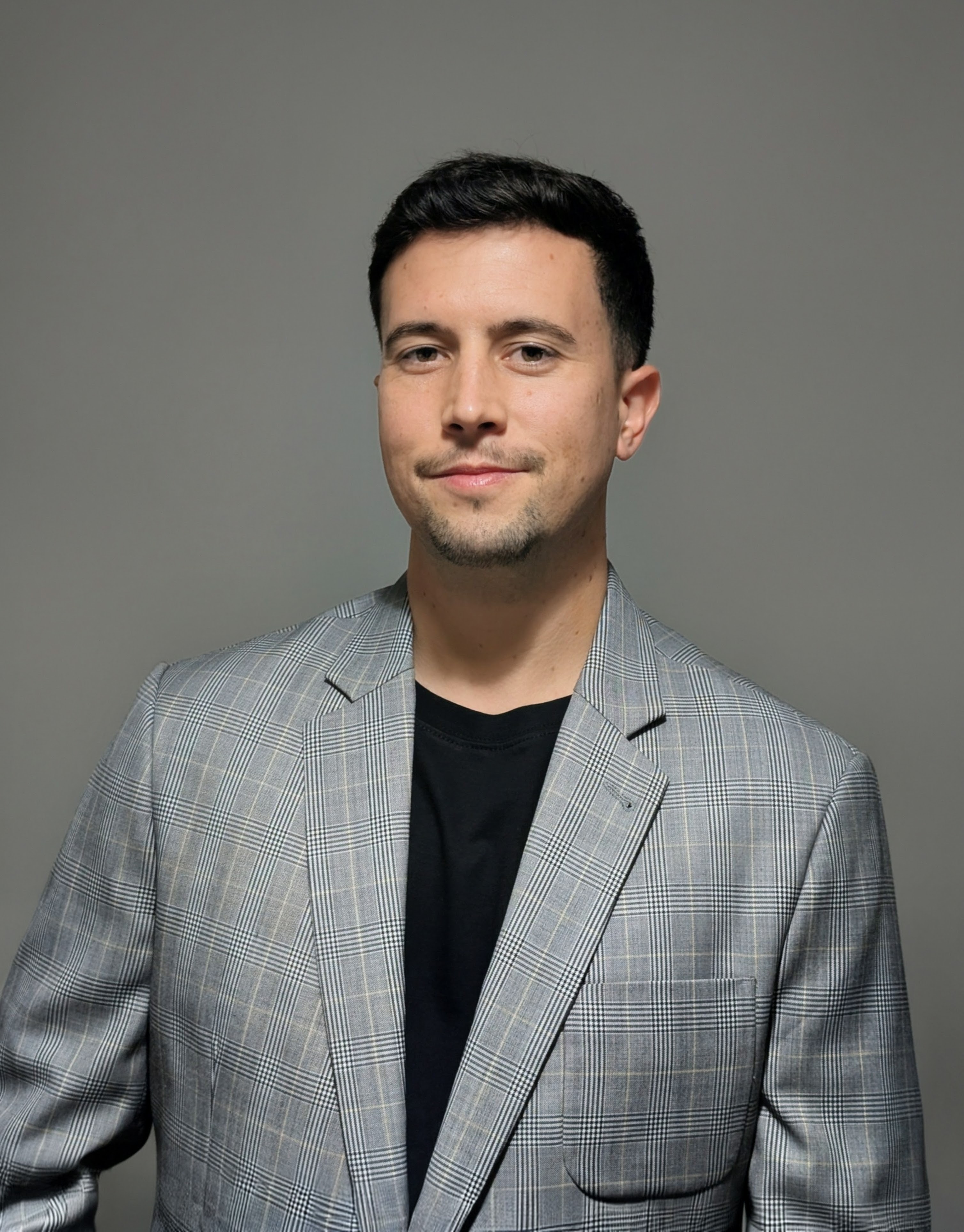 Dean is a white man with dark short hair, he is pictured wearing a black t-shirt and a gray checkered suit jacket.