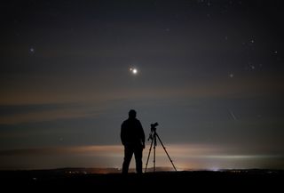 a silhouette of a person standing next to a camera and looking at two bright points of light in the sky, the larger brighter one is Jupiter and the smaller redder one is Mars. A long Perseid meteor streaks through the sky to the right.