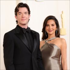 John Mulaney wears an all-black tuxedo while Olivia Munn wears a bronze dress while attending the Academy Awards