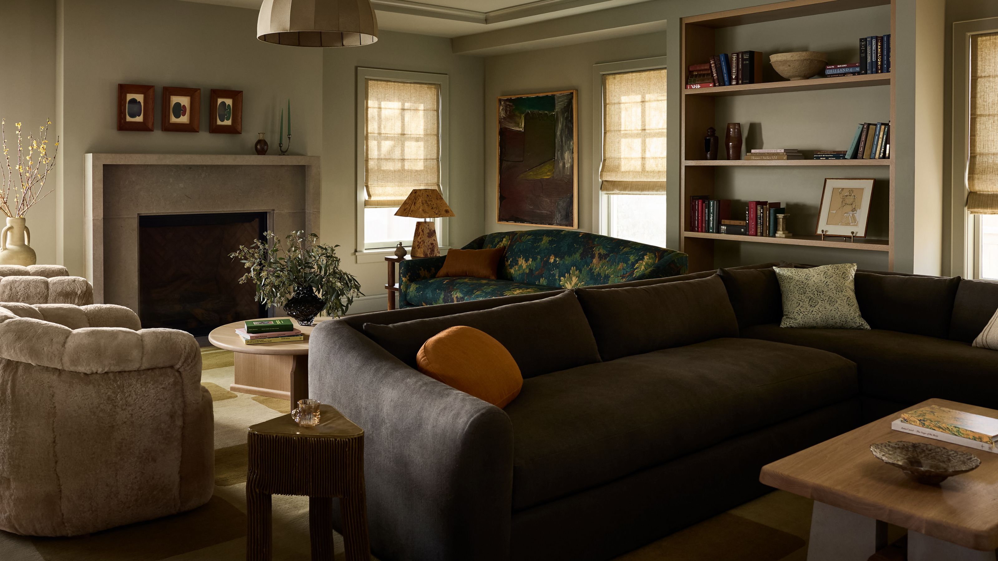 a muted green gray multi-purpose living room with a large chocolate brown couch, fireplace, shearling armchairs, tapestry couch and a bookcase