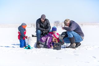 Three young children with their dad and grandpa ice fishing on a frozen lake in late winter early springtime The ice is covered with snow The two sisters are kneeling near Grandpa who is getting a fishing pole line ready for them to fish with Dad is sitting on the portable fish housesled while the young boy looks on