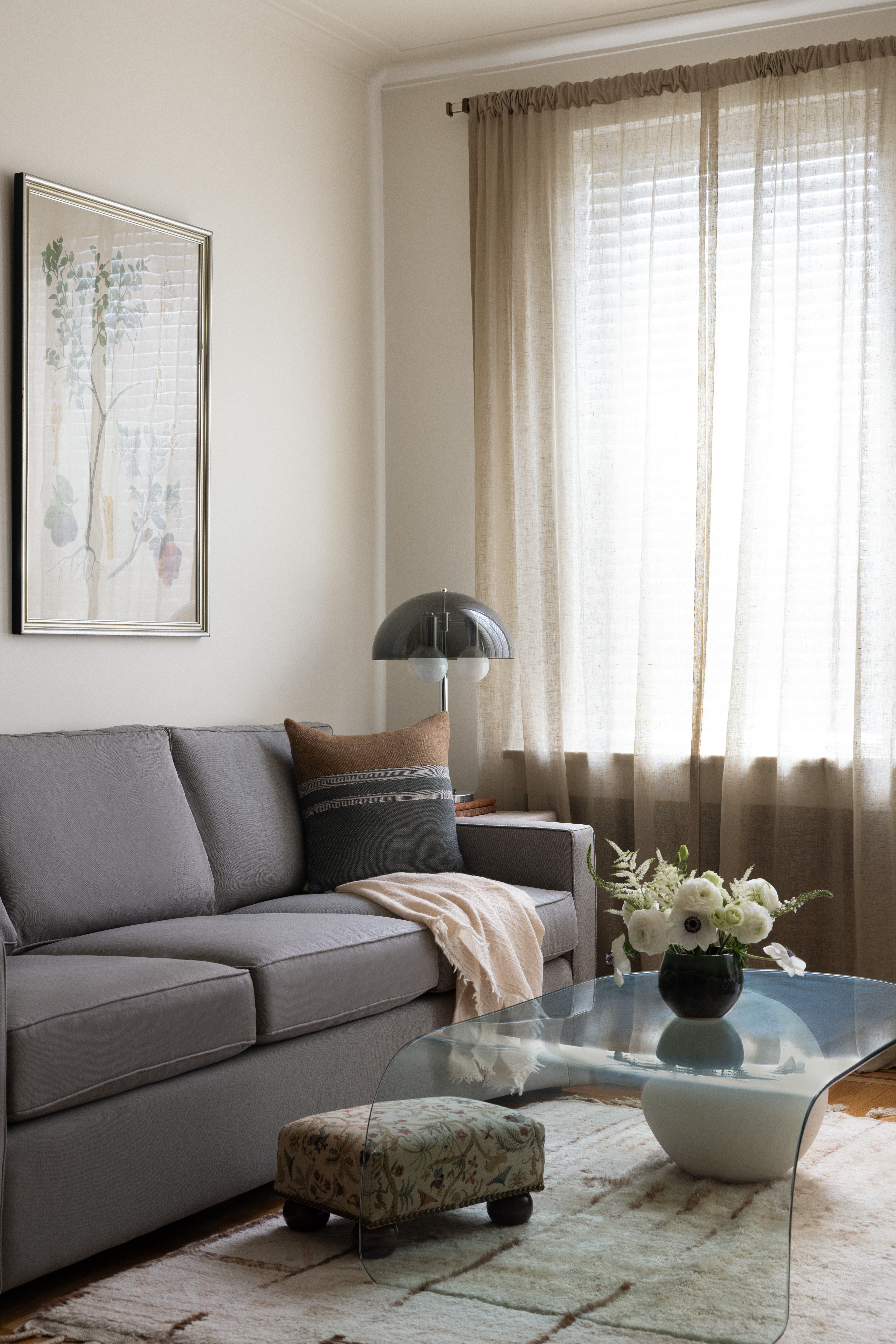 A apartment living room with a gray sofa, a mid-century modern lamp, a glass coffee table, and a patterned fabric foot stool.