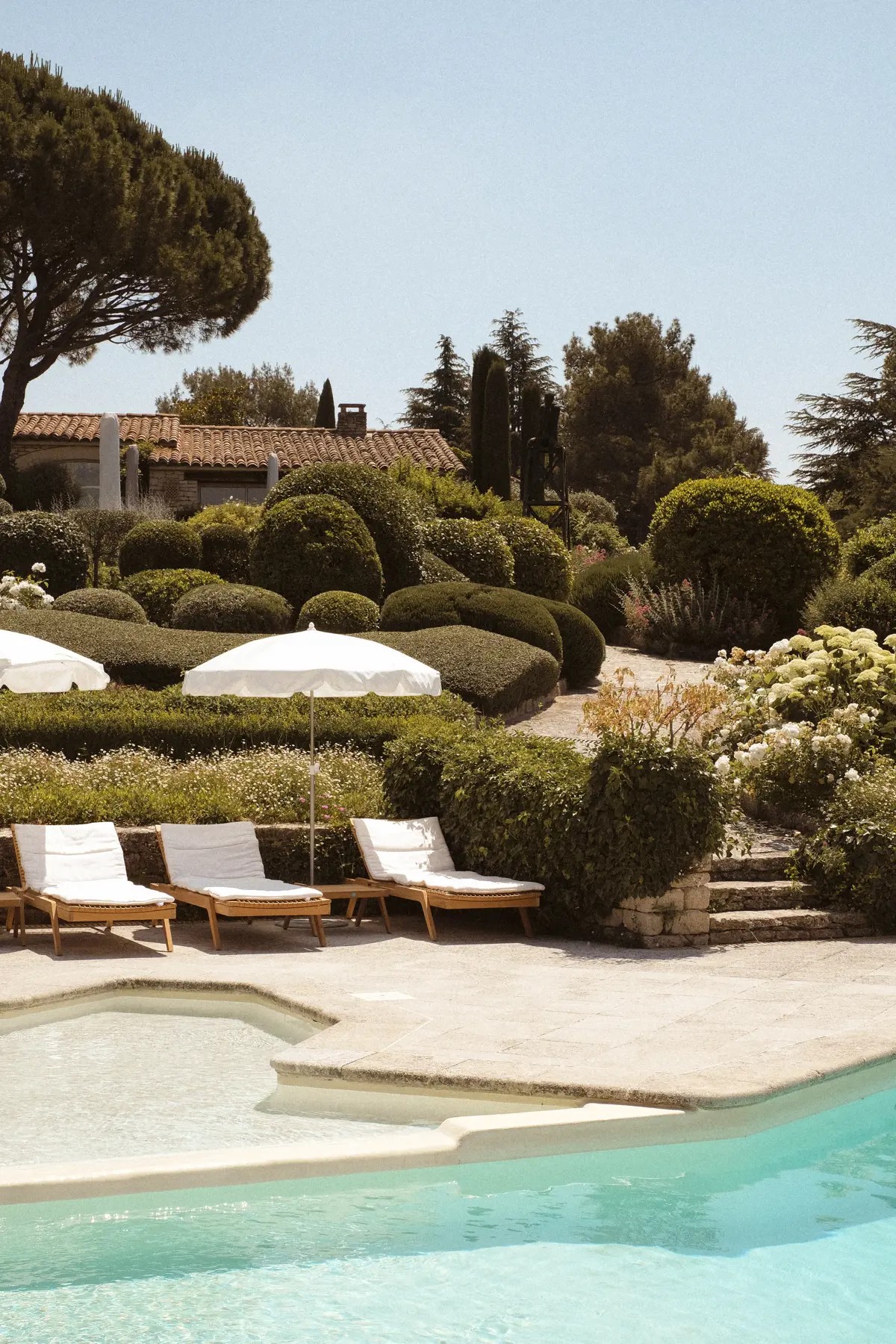 A sun-filled outdoor pool surrounded by reclining loungers in pale wood and white fabric and white parasols, alongside beautifully landscaped plants sprouting against raw rock.