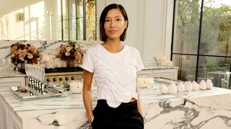 Imogen Kwok, a young chef and artist, stands in a sun-lit room wearing a wet-effect, white T-shirt and black trousers in front of a marble kitchen counter adorned with beautiful glasses, salt-crusted pears, and candles.