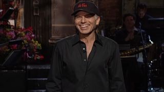 Billy Bob Thornton stands smiling on stage while wearing a baseball cap during his monologue for SNL S27 E6.