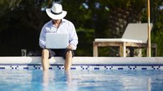 A man wearing a fedora and business suit, his pant legs rolled up, sits with his feet in the pool.