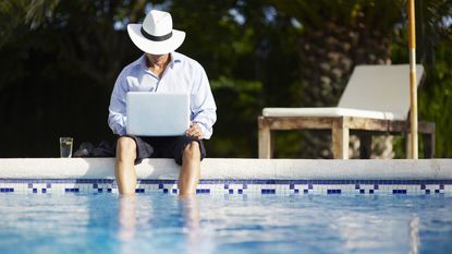 A man wearing a fedora and business suit, his pant legs rolled up, sits with his feet in the pool.