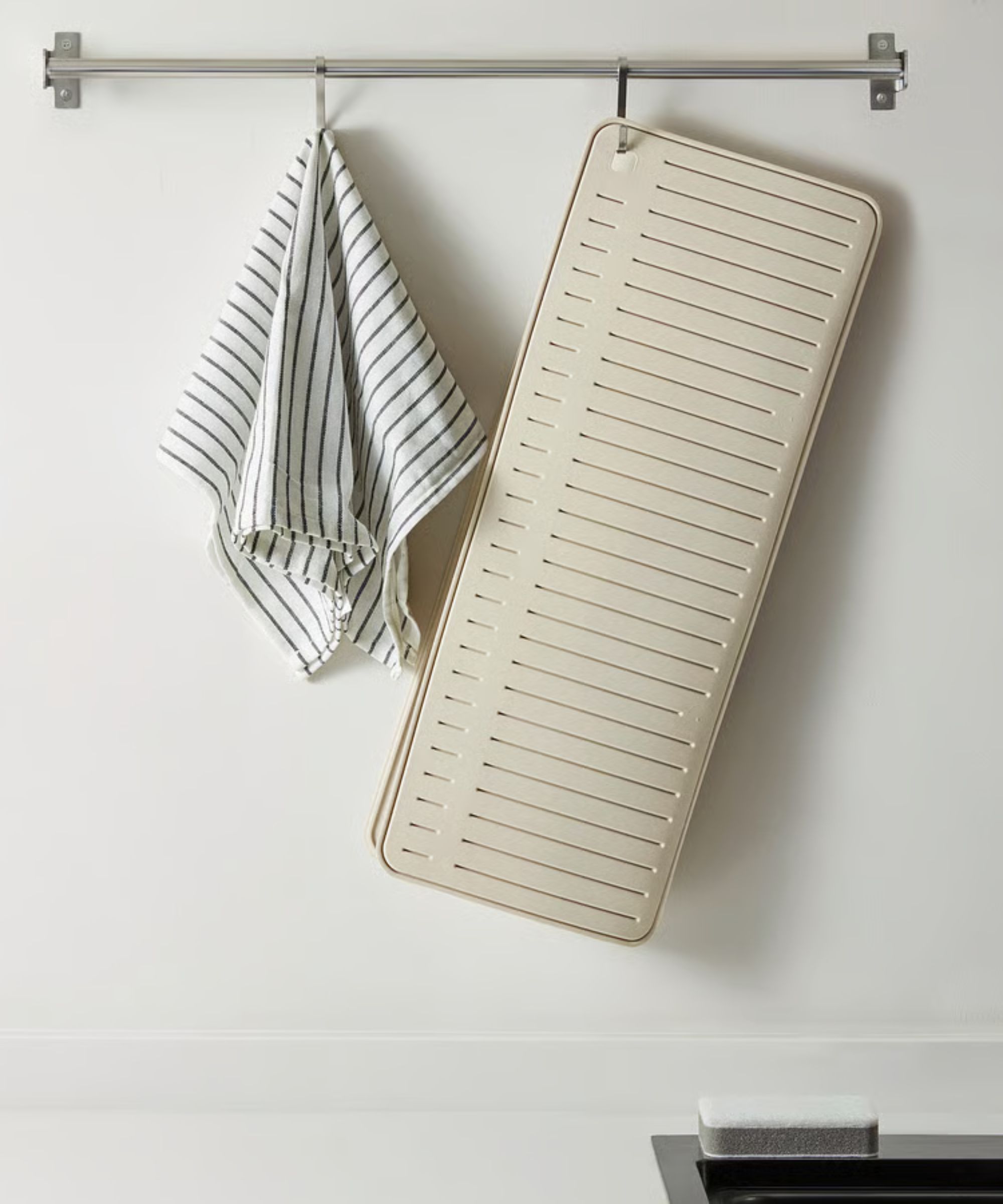 A beige dish drying mat hanging from a stainless steel hook against a white wall.