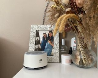 Dreamegg D1 white noise machine in white sitting on nightstand in front of photo in frame, beside skincare products and vase with dried flowers