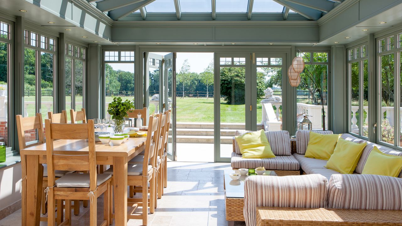 Green framed conservatory with wooden dining table and wicker u-shaped sofa