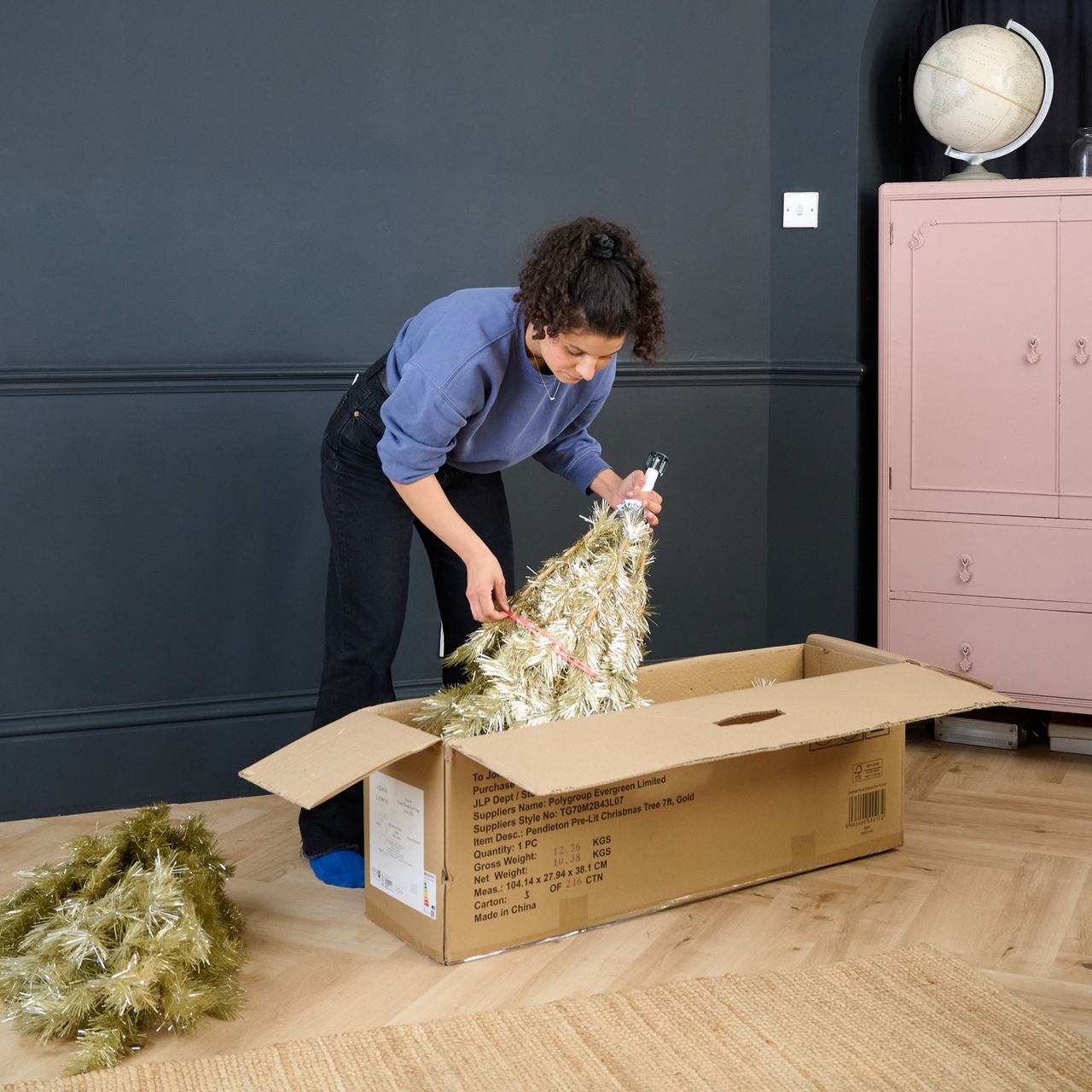 Sara taking out a John Lewis gold Christmas tree from its box in a black-painted living room