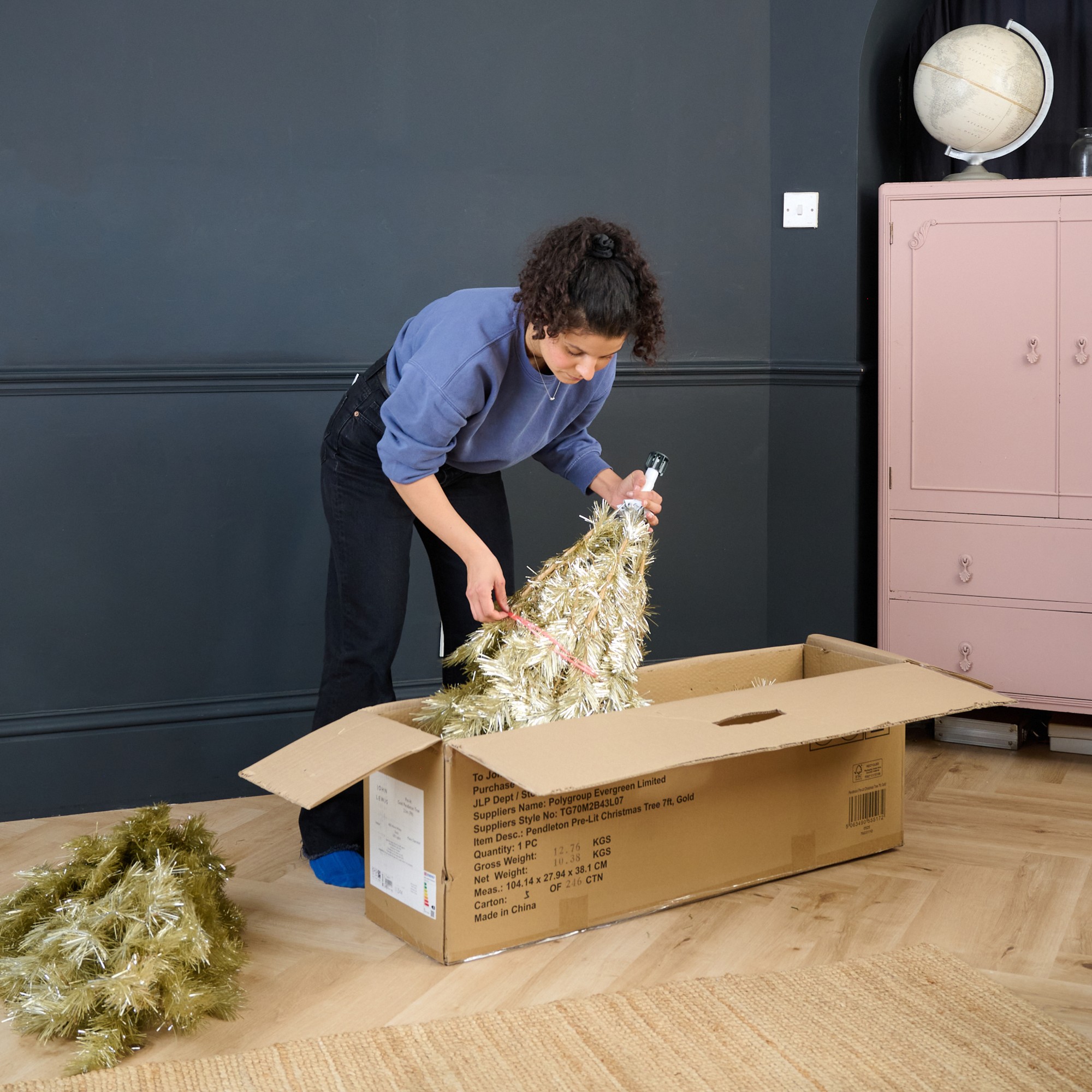 Sara taking out a John Lewis gold Christmas tree from its box in a black-painted living room