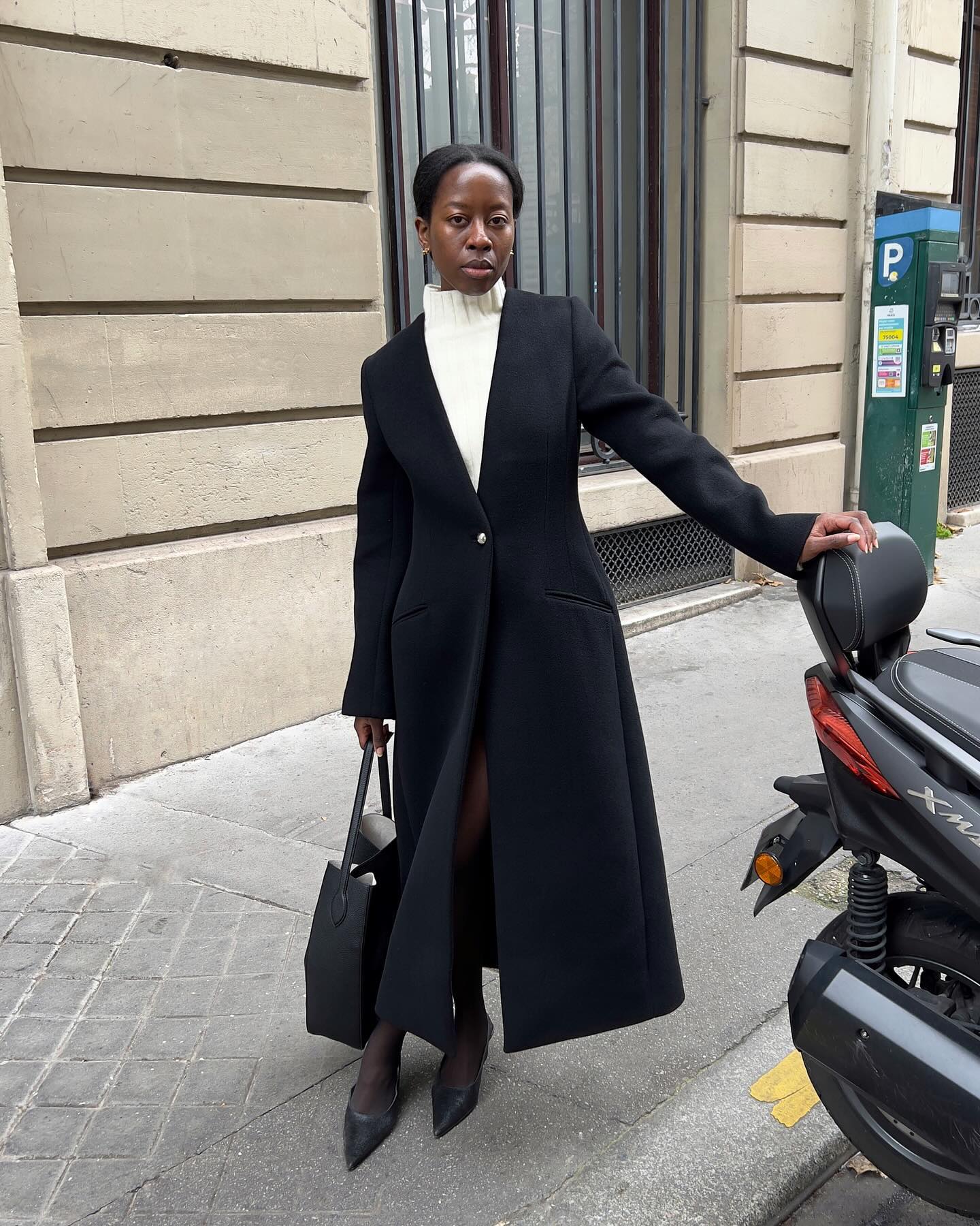 Sylvie wears a black cinched long coat, white turtleneck, tights, black heels, and a black tote bag.