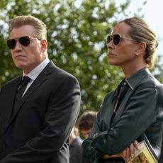 Holt McCallany and Maria Bello as the buckleys standing at a funeral with sunglasses on in the waterfront