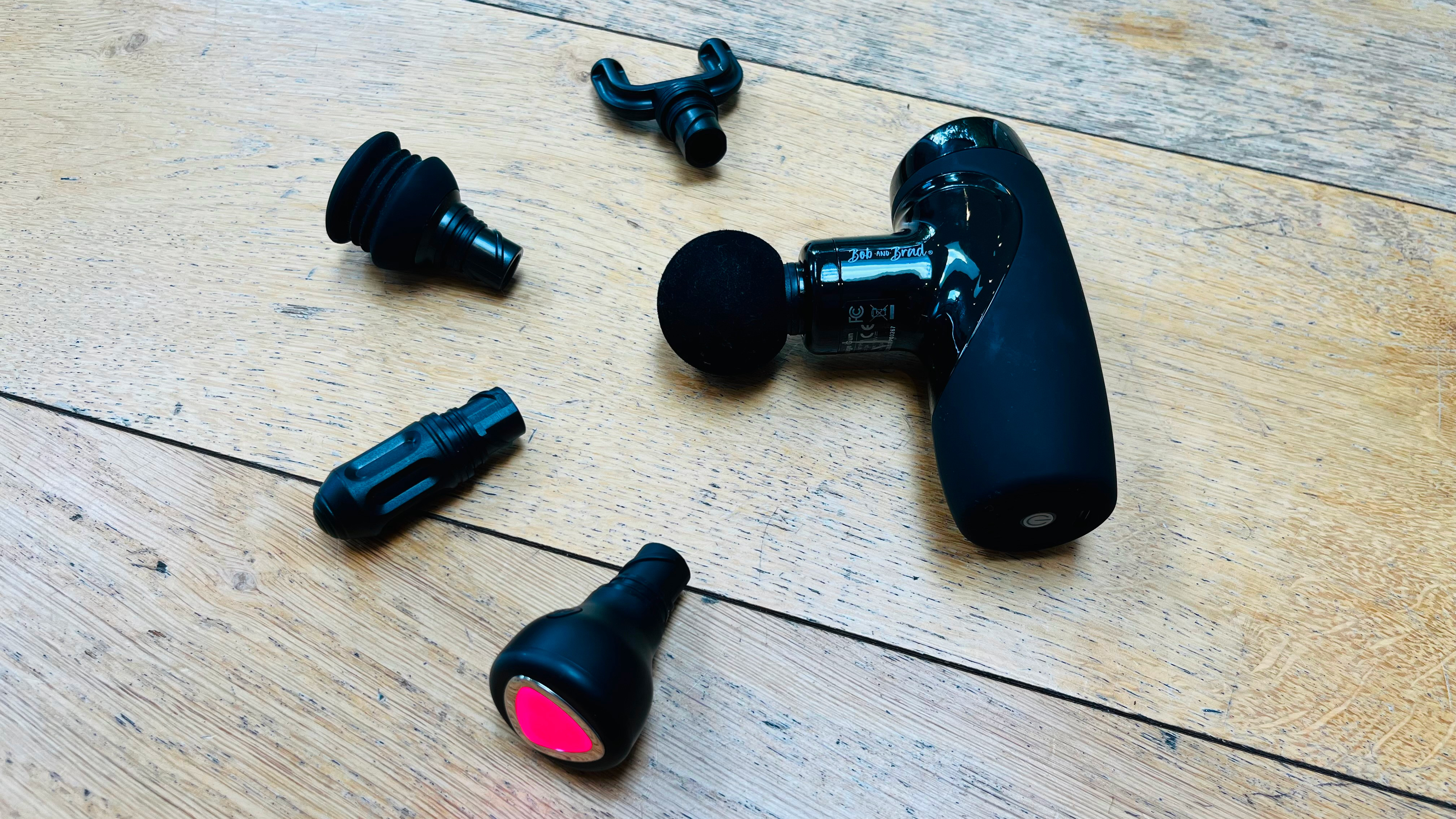 Massage gun lying on wooden floorboards with four head attachments arranged in a semicircle to the left of the massage gun