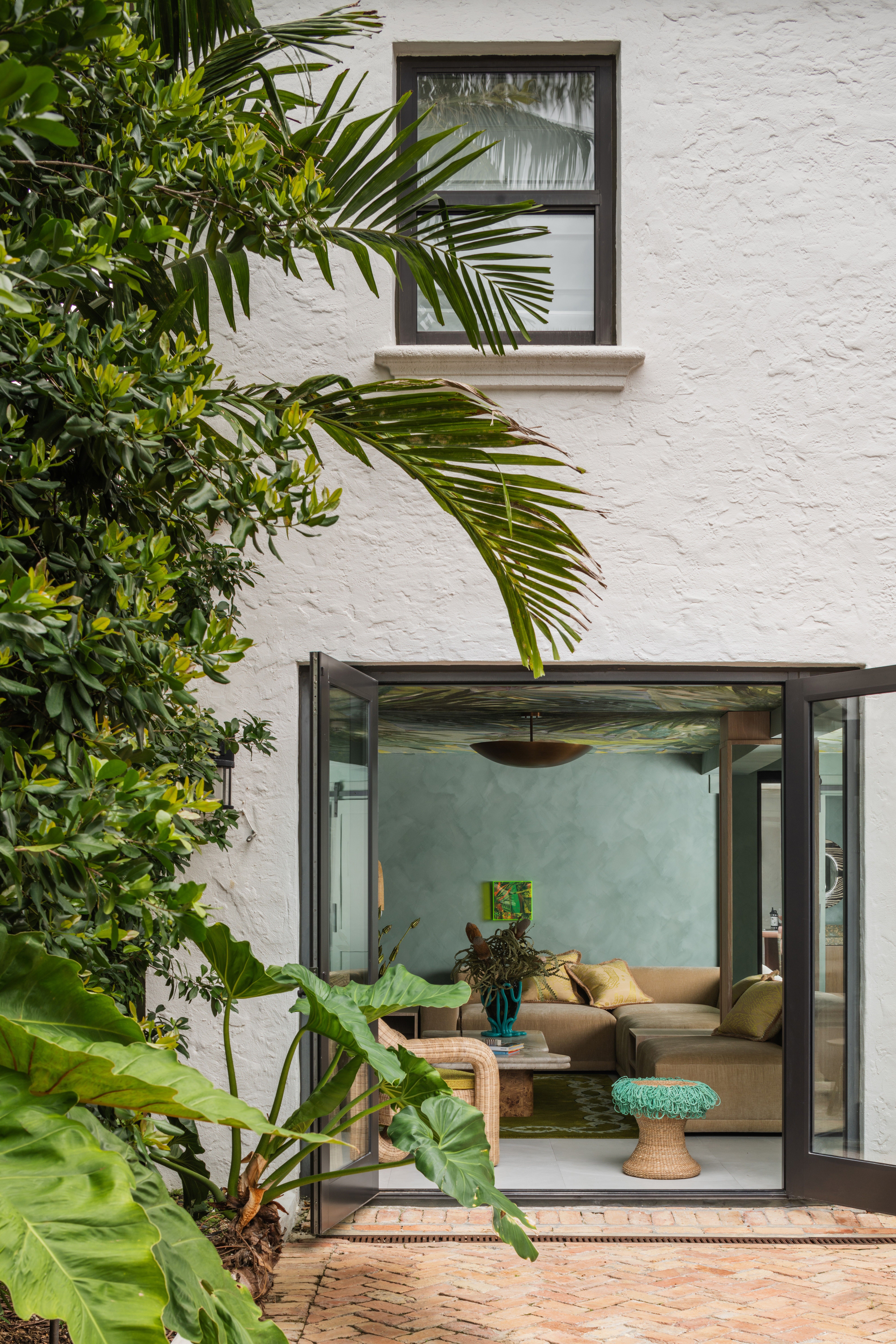 Interior of a tropical home with teal walls, zellige tiles surfaces, wavy, sand-coated details, rattan furniture mixed with bright aquamarine and ochre touches, and a botanical-themed mural ceiling.