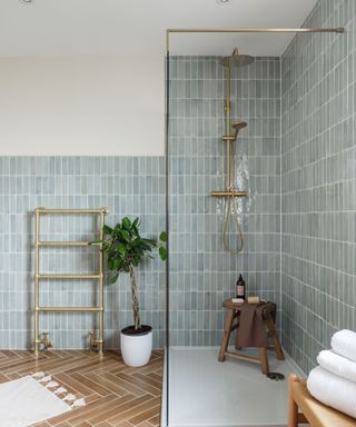 A small bathroom with a shower blue tiles with a gold shower head with a dark wooden stool underneath it, a gold towel rail and a white bath mat to the left