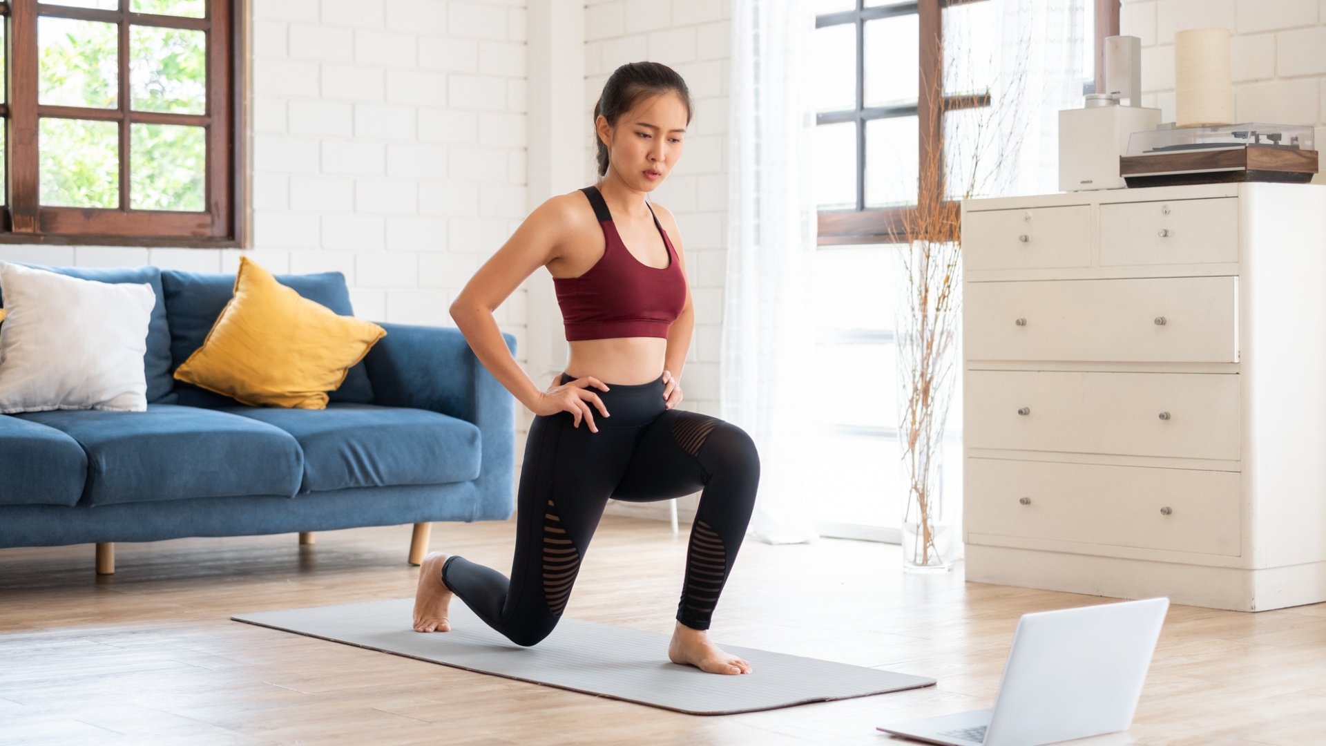 woman in black leggings and red crop top performing a lunge on an exercise mat in front of a laptop on wooden floor. there&#039;s a navy sofa and windows behind her and a white chest of drawers next to her.