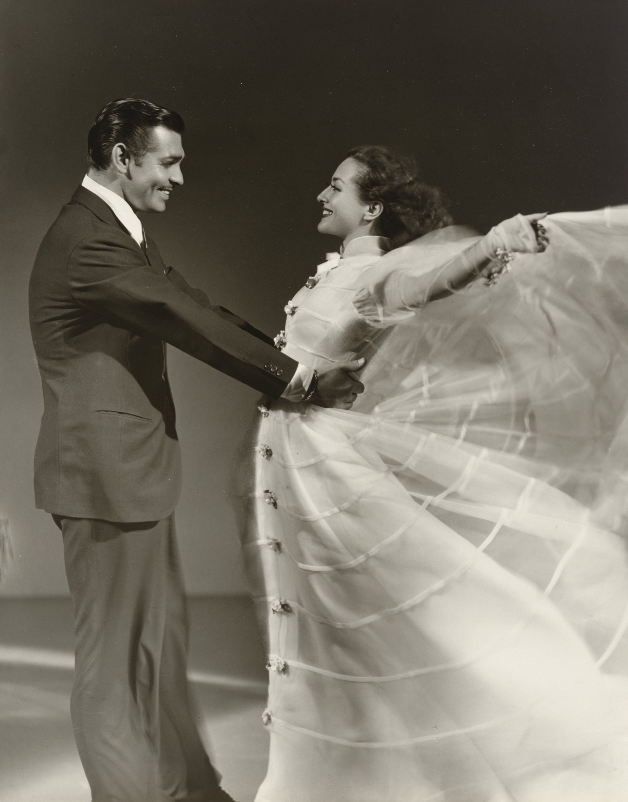 Clark Gable in a dark suit holds Joan Crawford by the waist as they smile at each other mid-dance, her sheer white dress and outstretched arms blurring into a sweeping fan of fabric.