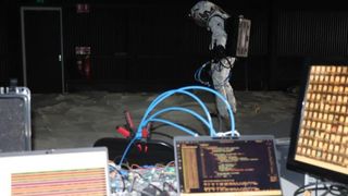 A person wearing a white suit stands behind a desk with various wires and computers on it
