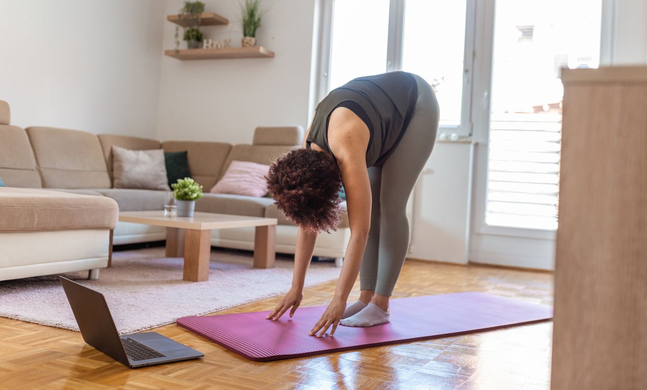 Woman in forward fold position in a living room setting. A laptop is on the floor in front of her. 