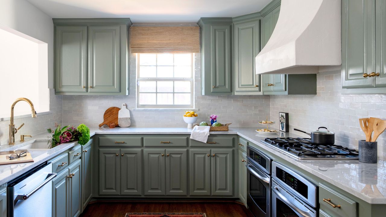 Narrow kitchen ideas are so pretty. Here is a kitchen with green cabinets and white marble countertops, a wooden floor with a red rug, a gold tap, a window with a woven blind, and an oven with a plaster hood over it