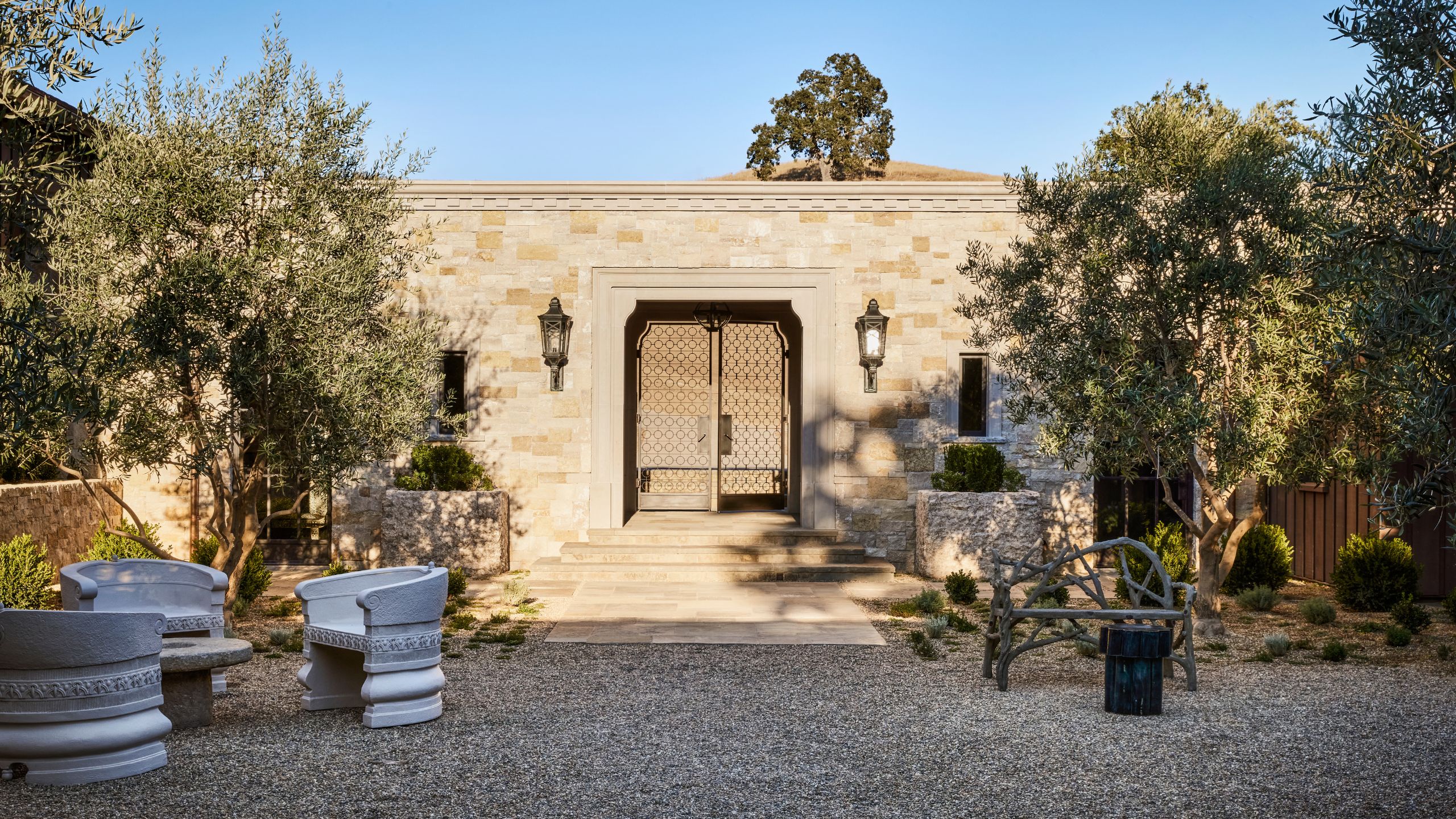 an exterior shot a large ranch-style home with ornate metalwork, trees, shingle, and carved courtyard furniture