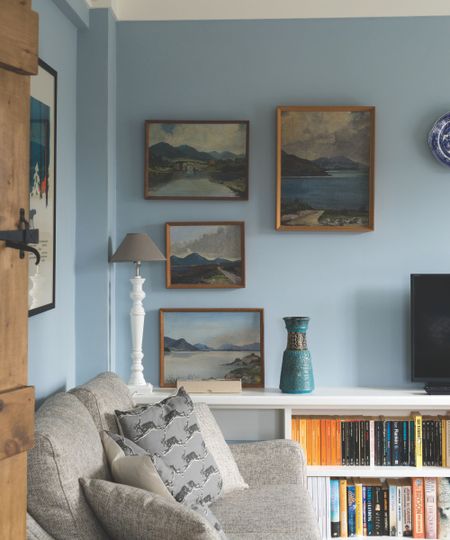 Living room with pale blue walls, gallery wall of landscapes, a grey sofa and cushions and a bookshelf with books