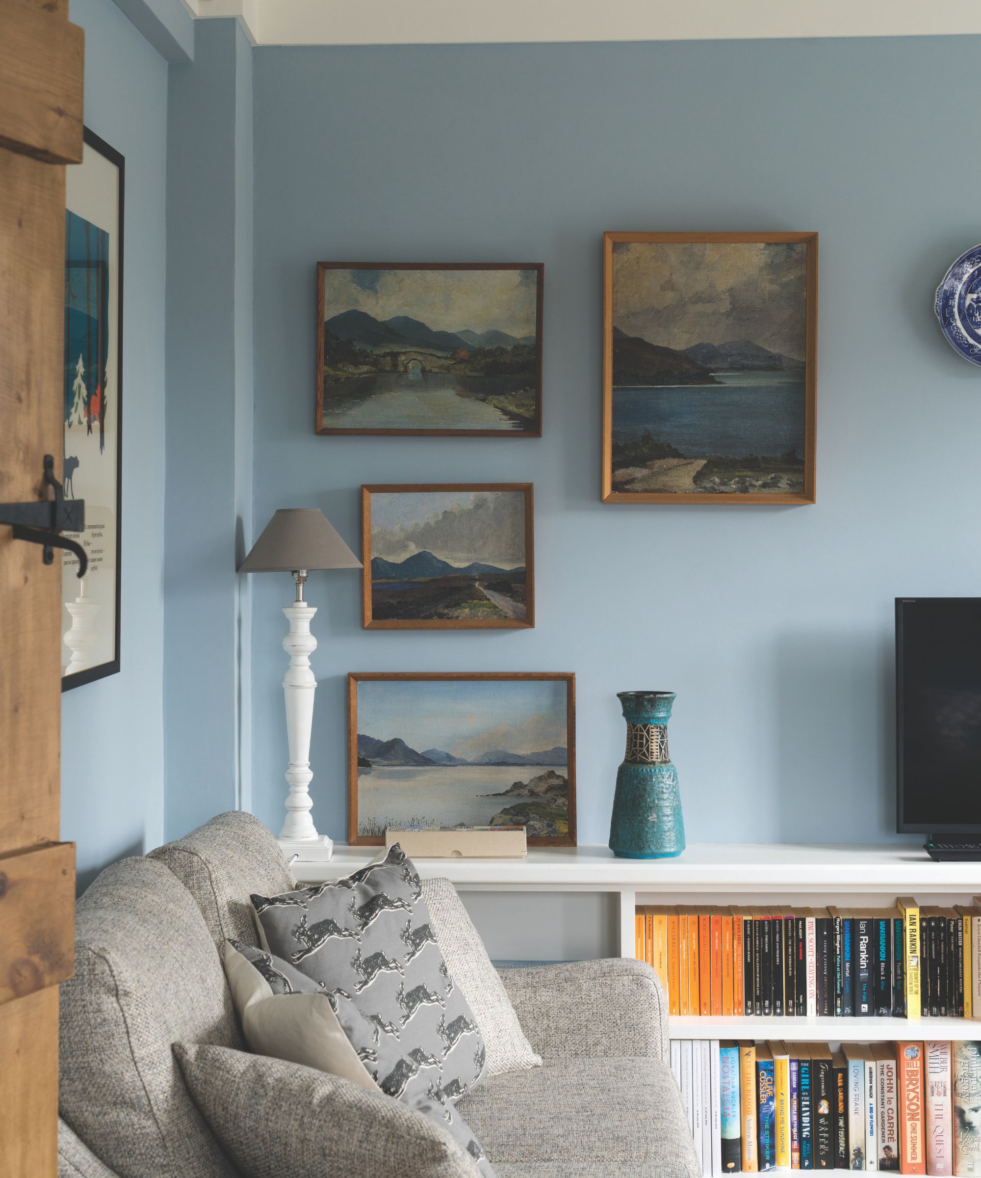 Living room with pale blue walls, gallery wall of landscapes, a grey sofa and cushions and a bookshelf with books