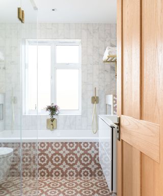 A light wooden door opening up to a bathroom with marble walls, a window, a white bath with brown and white circular tiles running on the edge of it and on the floor, and a glass shower