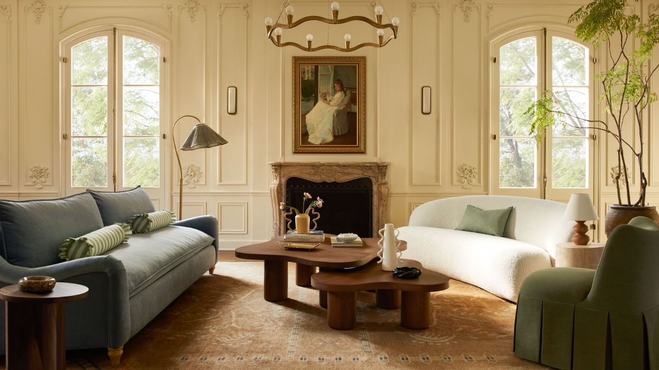 A blue sofa, a white sofa, and a blue pouffe surrounding a dark brown coffee table; in the background, an ornate fireplace and a portrait of a woman hanging on white walls with windows and a garden view. 