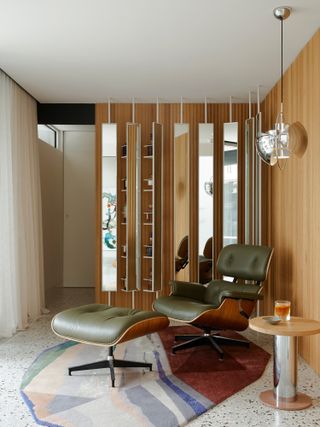 A room with olive green armchair and footstool and mirrored panels concealing storage for a home bar