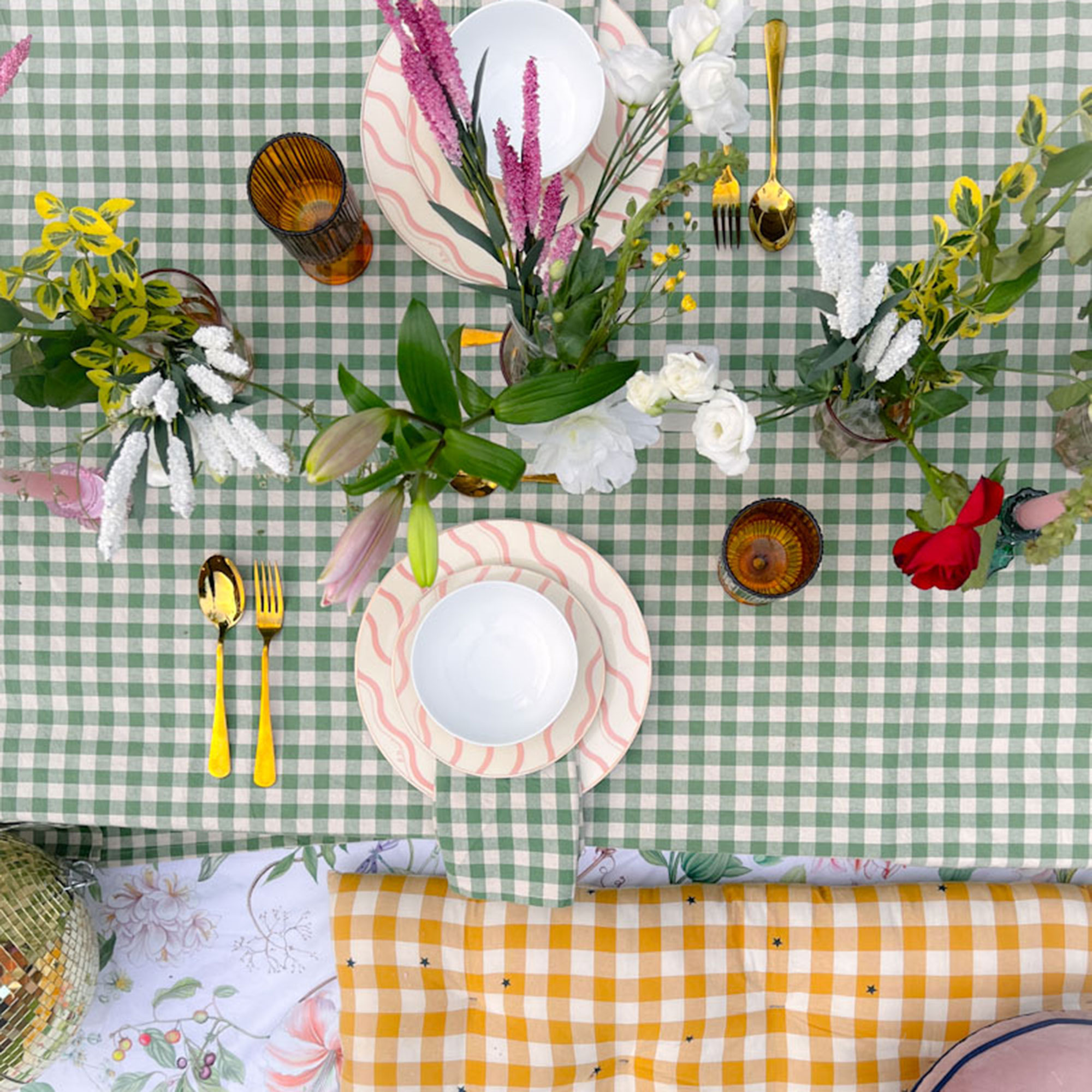 Overhead view of table decorated with green and white gingham tablecloth, flowers and foliage, gold cutlery and flatware with pink and white squiggle pattern