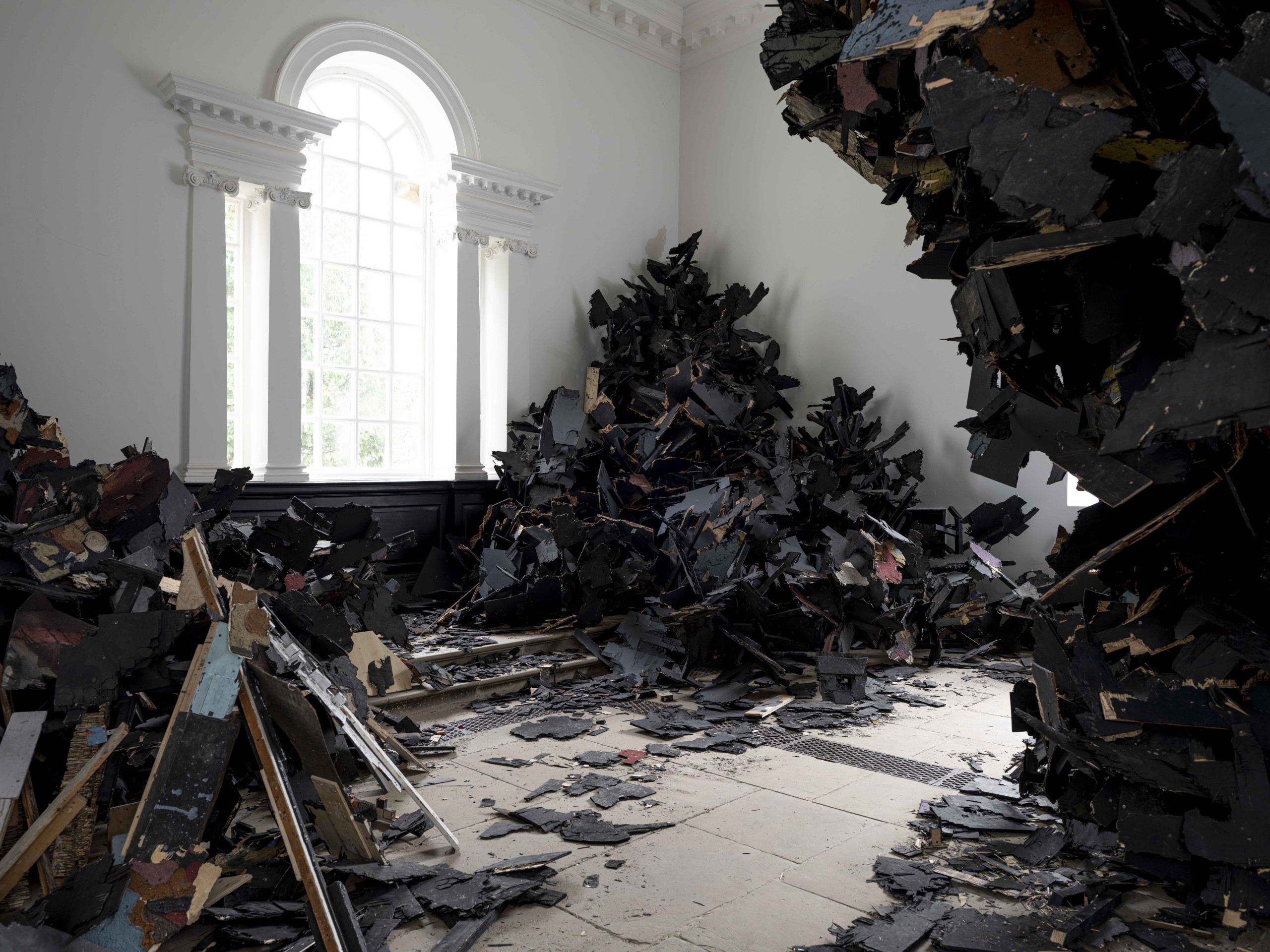 A large-scale art installation made of burned up debris occupies a palatial gallery room with stuccoed detailing and grey floors.