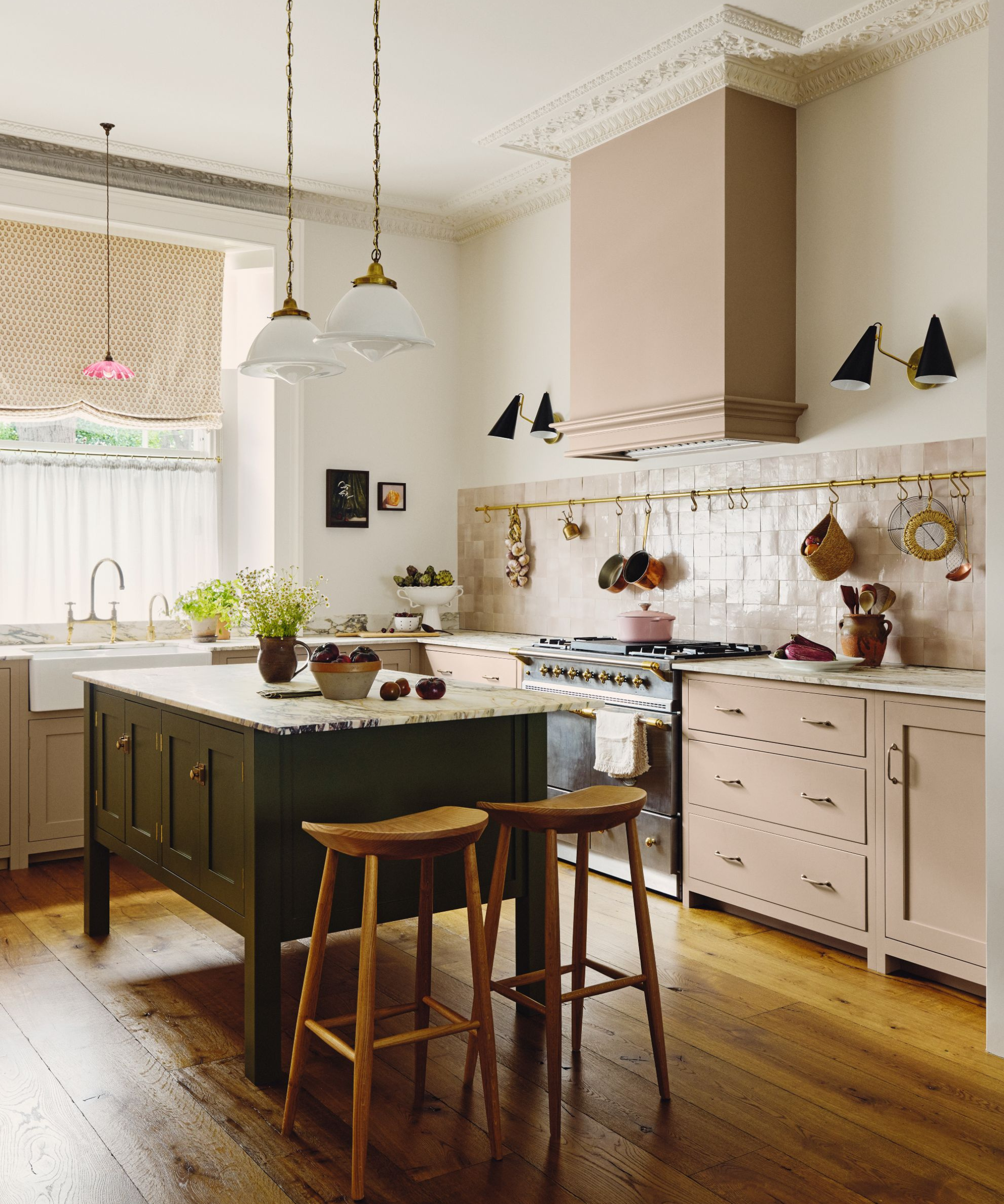 a kitchen with dusty pink cabinets and backsplash, an island with a green base and a marble countertop, pendant lights, and a rack of pots and pans hanging against the wall
