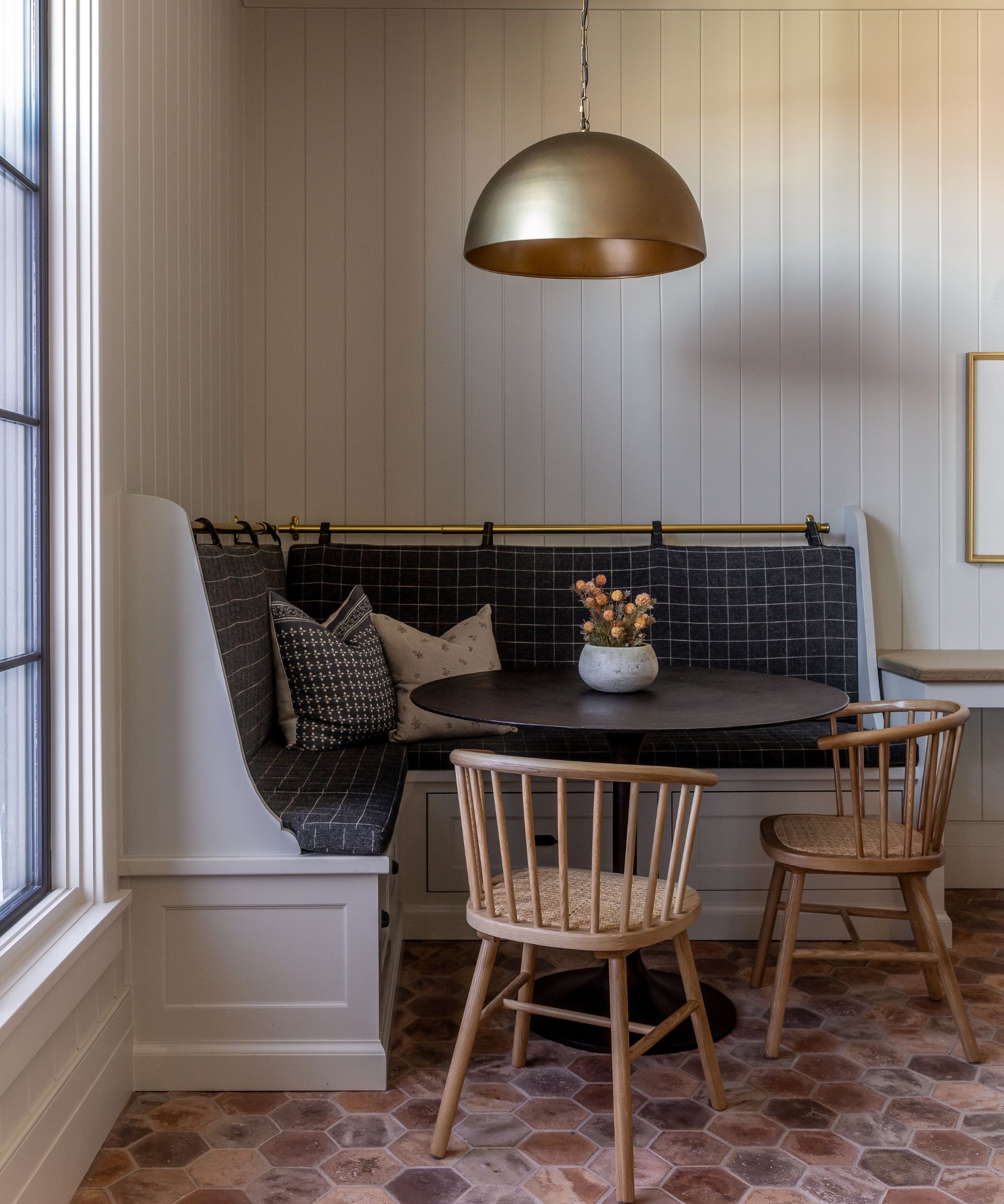 A cozy breakfast nook with taupe wood-clad walls, banquette seating with a dark gray grid-pattern cushion, a small round table with two wooden chairs, and terracotta tile flooring.