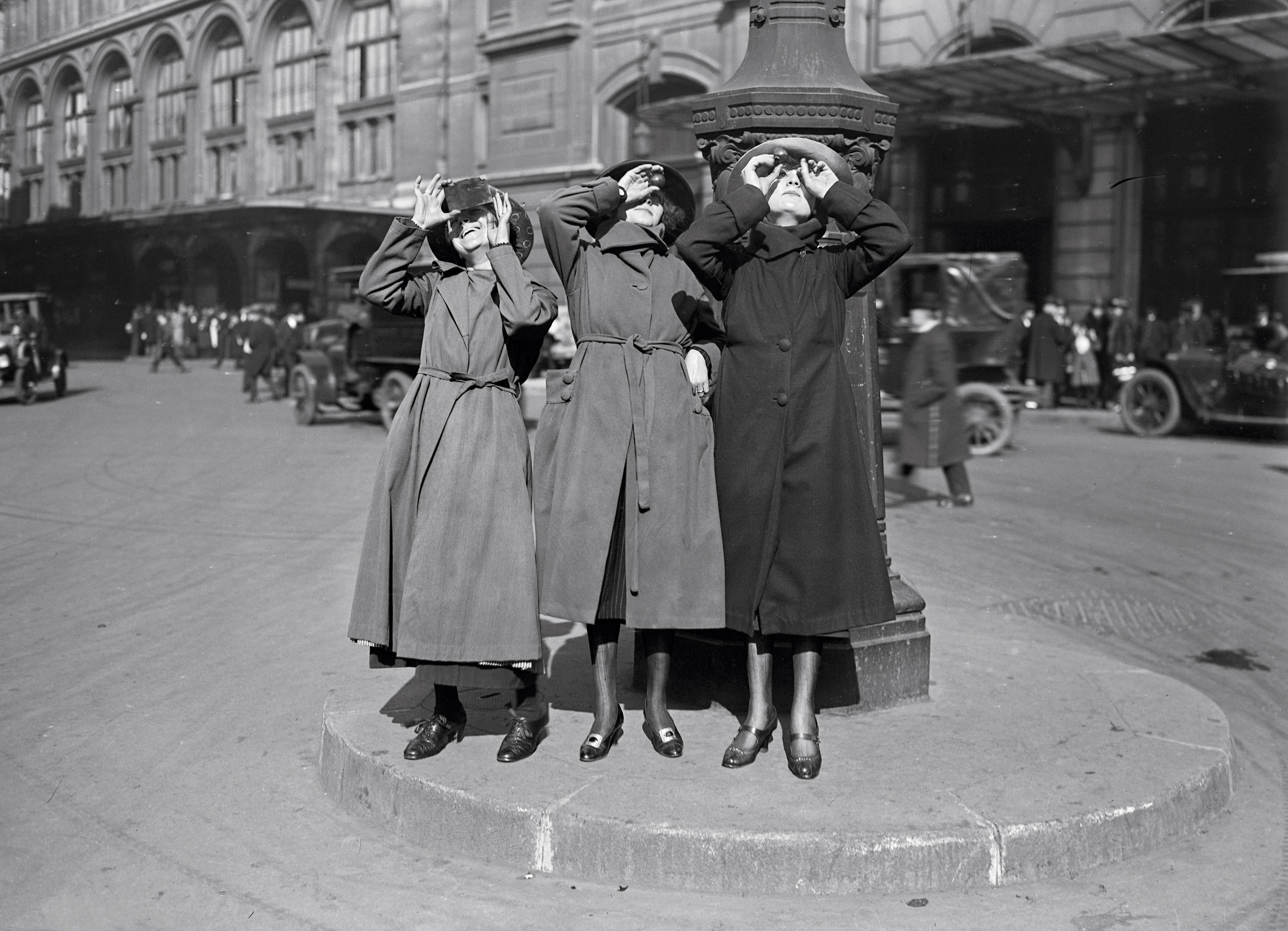 three women wearing long coats look up at the sky with photographic film.