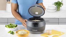One of the best rice cookers on the market, the Nutribullet EveryGrain rice cooker on the countertop with a bowl of rice and a bowl of cilantro around it. A man in a blue top is lifting the lid of the rice cooker to show some white rice inside